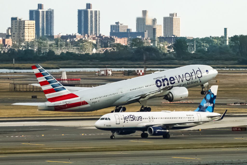 An American Airlines Boeing 777 in special Oneworld livery takes off from New York JFK with a JetBlue Airbus A320 taxiing in the foreground