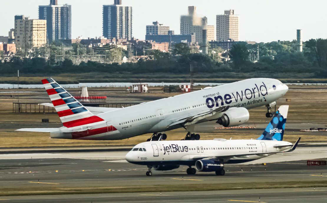 An American Airlines Boeing 777 in special Oneworld livery takes off from New York JFK with a JetBlue Airbus A320 taxiing in the foreground